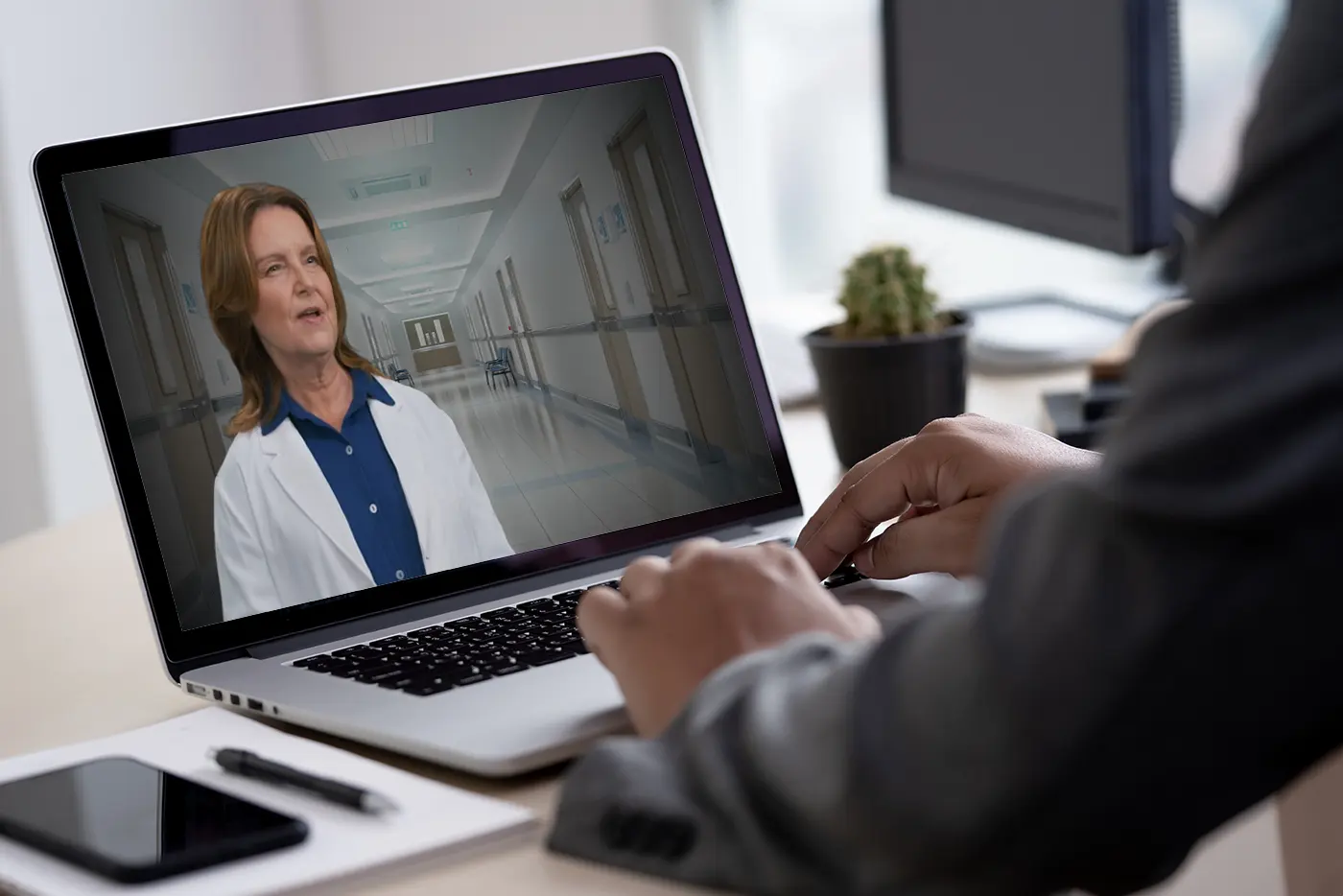 Over-the-shoulder view of a person at a laptop watching a video of a clinician in a white coat standing in a hospital corridor